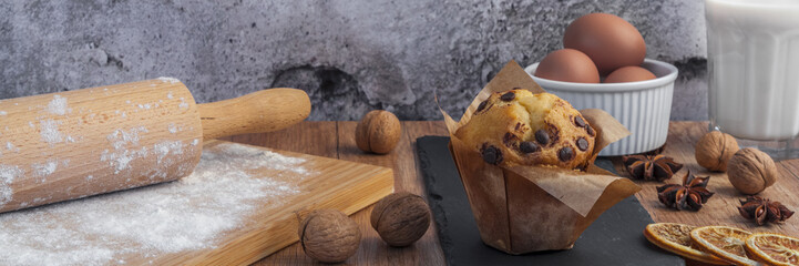 Panorama with a chocolate muffin, a glass of milk, organic eggs in a white bowl, rolling pin, flour sprinkled on a wooden board, dried oranges, walnuts, anise and a stone plate on a brown wooden table