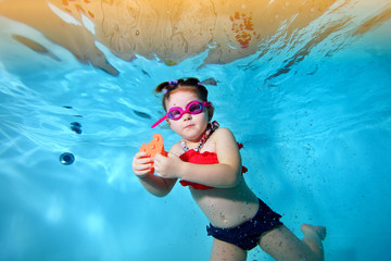 Beautiful little girl swims underwater in the pool with a toy in his hands and looks at me....