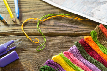 The process of cross-stitch. Needles, floss, scheme, scissors, pencils and pen on a wooden table.