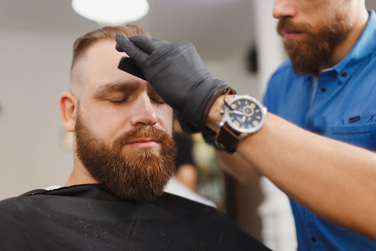 Male professional hairdresser serving client, shearing eyebrows with scissors. Ginger stylish young man with thick big beard and short hair getting trendy haircut in black cape. Light barber shop room