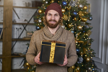 Fototapeta premium Young man cheerful in a sweater gift in a hand Christmas tree on background, joy, happiness, new year