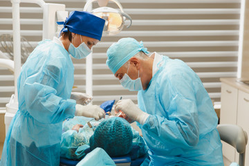 Male old professional dentist surgeon in uniform and female assistant helps to performing operation install dental implant teeth of woman patient in clinic light office with modern tools equipment.