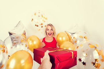 Beautiful blond woman in red t-shirt receives a gift in red packing and gold balloons from man on a white background