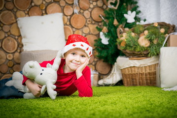 boy waiting for a holiday in a room decorated before Christmas. boy in a red sweater sits on a green carpet. cute boy in santa hat
