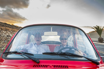 Obraz premium Elderly couple with hat, with glasses, with gray and white hair, with casual shirt, on vintage red car on vacation enjoying time and life. With a cheerful mobile phone smiling