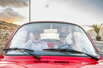 Fototapeta na wymiar Elderly couple with hat, with glasses, with gray and white hair, with casual shirt, on vintage red car on vacation enjoying time and life. With a cheerful mobile phone smiling