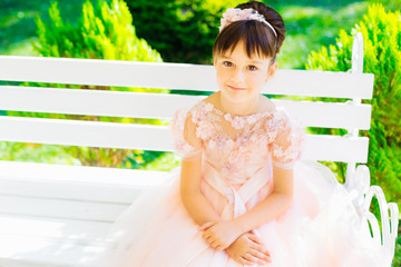 Young girl in a pink dress sitting on a white bench on a background of green bushes