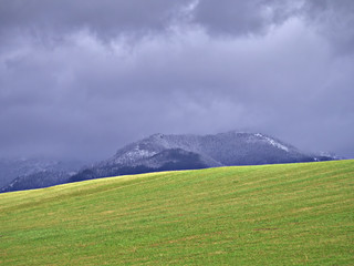 Heavy snowfall at mountains behind green field. Late autumn seasonal background.