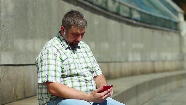 Man uses smartphone and listens to music. Businessman sits on the step and listens to music on his smartphone. Fat guy with red mobile phone