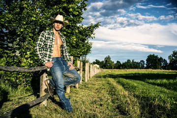 Portrait of sexy farmer or cowboy in hat with unbuttoned shirt on muscular torso, looking to a...