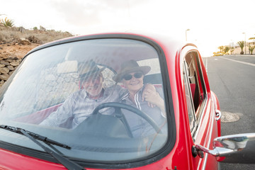 Elderly couple with hat, with glasses, with gray and white hair, with casual shirt, on vintage red car on vacation enjoying time and life. With a cheerful mobile phone smiling 