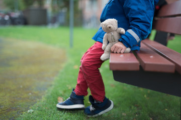 boy sitting alone on the bench in a park with his bear