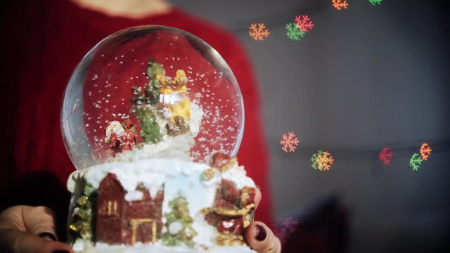 clouseup hands holding magic globe with snow and santa, on background colorful garland with snowflake figure bokeh