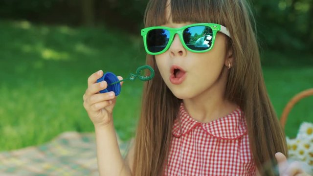 Portrait of the funny small girl in kids' green glasses blowing soap bubbles in the middle of the green park. Close up. Outside