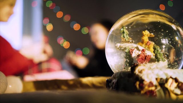 clouseup magic globe with snow and santa, on background blurred figure mother and daughter on the bed and garland in bokeh