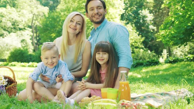 Young cheerful parents having a picnic with their small pretty daughter and son in the picturesque green garden on a sunny day of summer. Portrait shot. Outdoors