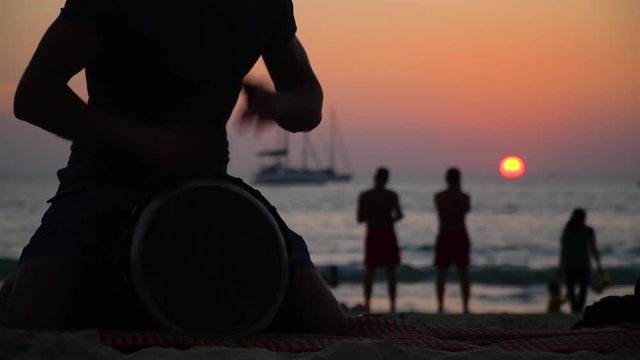 A man is playing on a djembe drum on the beach at sunset. Against the background are people admiring the sea sunset.