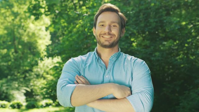 Portrait of the attractive Caucasian man with a beard posing to the camera, looking straight and crossing his hands in front of him. Beautiful green park. Close up. Outdoors