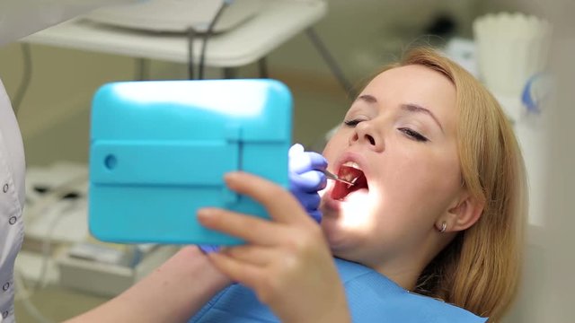 A young girl looks in the mirror on examination at the dentist. Doctor dentist tells and shows the girl on her problem in the teeth in the mirror.