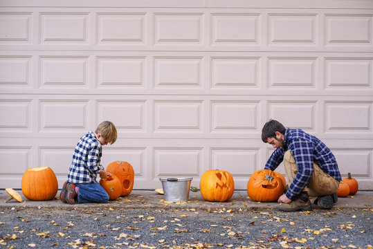 Father and son sitting by a garage door carving Halloween pumpkins