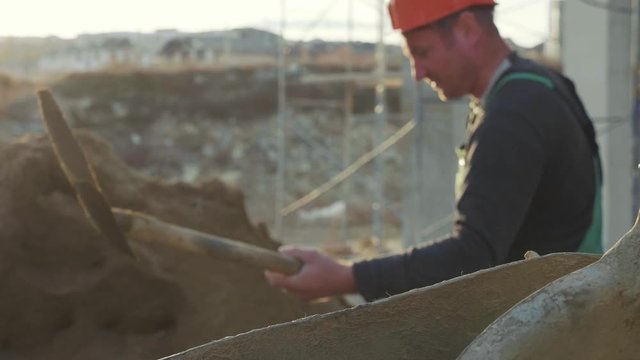 Caucasian worker pours sand into a concrete mixer by shovel at construction site