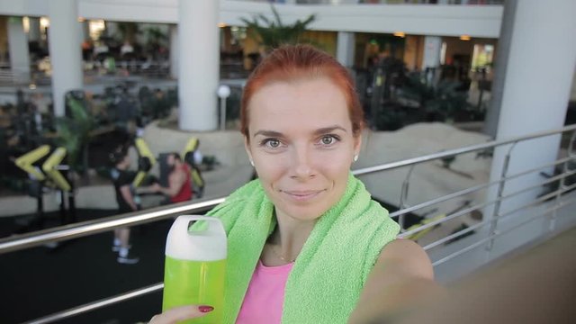 The portrait of young woman doing selfie on training in the modern gym. The sportswoman with red hairs wears pink top and has green towel on her shoulders. The athlete holds the bottle by her hand and
