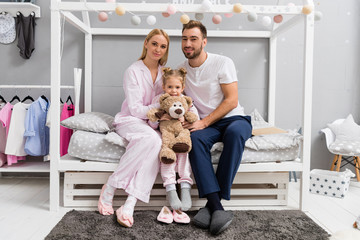 happy young family sitting on bed in kid bedroom and looking at camera