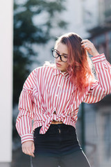 Portrait of young beautiful girl wearing stylish shirt, black skinny jeans, glasses. Girl have shinny long red hair. Female fashion concept