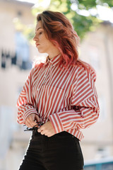 Portrait of young beautiful girl wearing stylish shirt, black skinny jeans, glasses. Girl have shinny long red hair. Female fashion concept