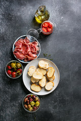 Ingredients for making tapas or bruschetta. Crusty bread, ham prosciutto, sun dried tomatoes, olive oil, olives, pepper, greens on plates over dark texture background. Top view with space © Natasha Breen