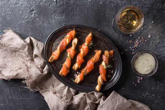 Appetizer with smoked salmon on crispy breadsticks served on vintage metal tray with cheese dill sauce, sea salt, glass of white wine, textile napkin over black texture background. Top view with space