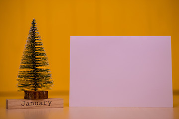 Word "January" on wodden table with christmas tree and blank white sheet of paper on yellow background. Christmas Holiday Composition with space for text