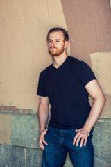 Fototapeta premium Moody Summer. Tough guy missing you. Wearing black v neck T shirt, jeans, a young man with beard, mustache leaning against painted wall, relaxing, sad, thinking, lost in thought. Man Street Fashion.