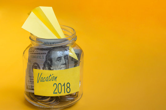 Coins, paper airplane and dollars in glass jar with vacation 2018 label on orange background. Saving money for travel, planning holiday or vacation. Copy space