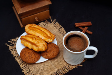 A cup of coffee, eclairs and oatmeal cookies on a black textured background.