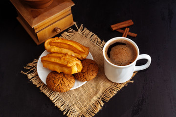 A cup of coffee, eclairs and oatmeal cookies on a black textured background.