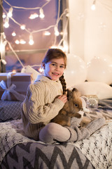 Very beautiful and charming little dark-haired girl in white sweater holds a live rabbit in the interior of the house. New year. Christmas. Hare. Studio.