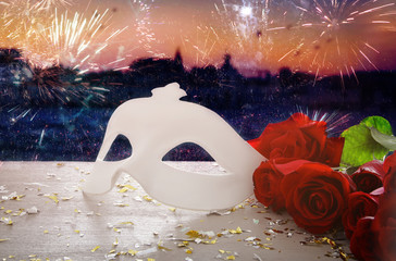 Image of elegant venetian mask and red roses over wooden table in front of blurry Venice background.