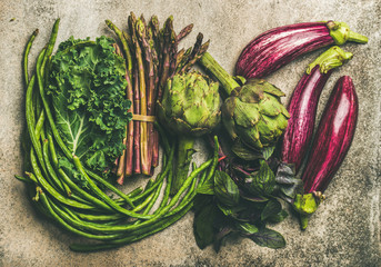 Flat-lay veriety of green and purple vegetables over grey concrete background, top view. Local seasonal produce for healthy cooking. Eggplans, beans, kale, asparagus, artichoke, basil. Clean eating