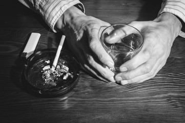 Hands of alcoholic man holding a glass with alcohol drink with smoking cigarette in the ashtray...