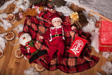 A child in Santa Claus costume rests on a red plaid surrounded by toys