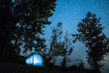 Blue tent set among trees on the coast of a river and night starry sky