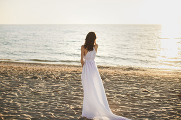 Fototapeta na wymiar Beautiful girl bride in a white dress and sneakers, at sunset walks along the beach.