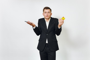 Young sad concerned disquieted business man in black suit holding golden credit card, mobile phone isolated on white background for advertising. Concept of money, achievement and wealth in 25-30 years