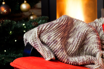 Fototapeta na wymiar warm knitted scarf on chair, christmas tree and burning fireplace in the background - hygge concept