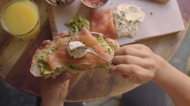 top view of woman that prepares sandwich or toast from toasted bread, avocado spread, smoked samon and creme fraiche with herbs. Gives it bite and enjoys tasty brunch or breakfast