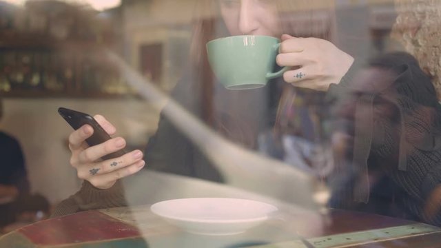 Through glass cinematic beautiful shot of amazing young natural beauty woman, that drinks hot chocolate or cacao drink, looks at smartphone, millennial always connected generation