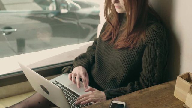Beautiful young woman chats or texts, surfs internet websites on aptop, while sits in downtown coworking startup hub or cafe. Smiles and enjoys futuristic technology that permits her to work remotely