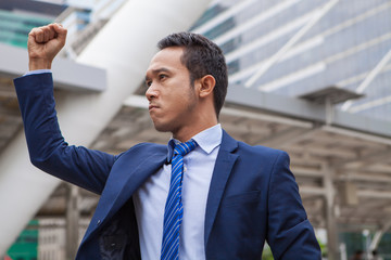 Businessman seriously  and raising his fist in the air, with office building background - business success, achievement, and win concepts