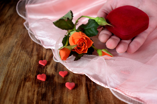 Male hand is holding a box for a ring. An orange rose and little hearts are lying next to it